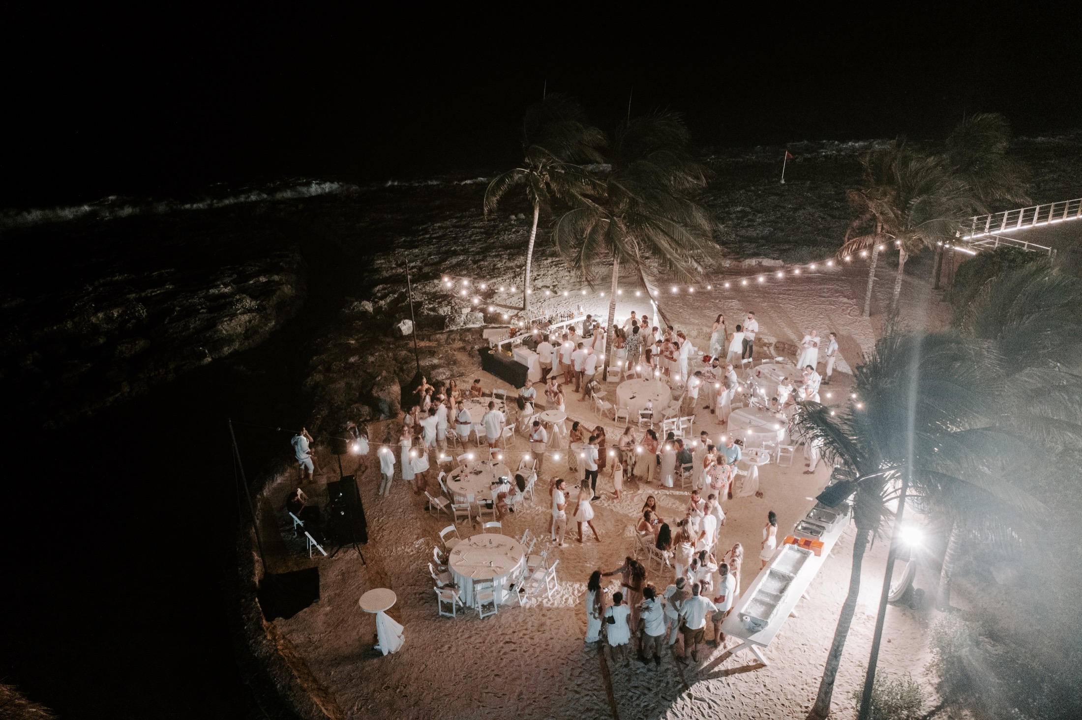 Beach wedding reception beneath string lights, seen from above